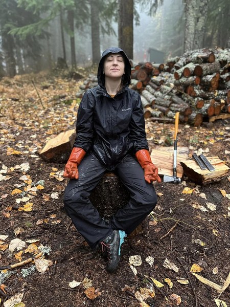 Woman in rain gear sits on stump in zen-like pose with axe visible behind her.